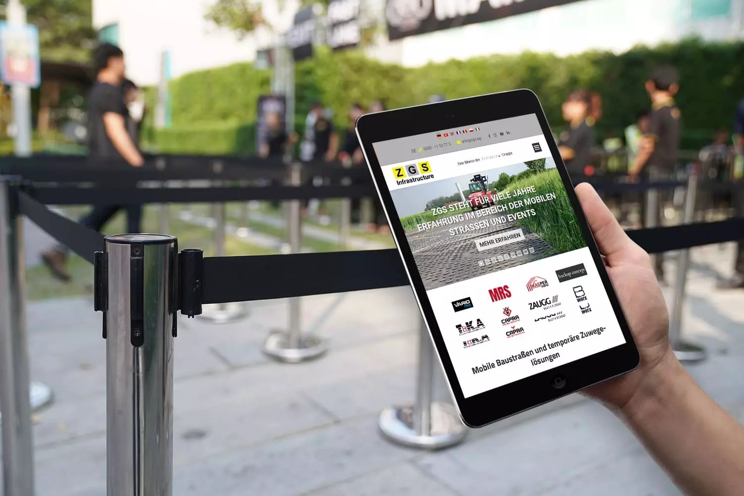 Person hält ein Tablet mit der Website von ZGS Infrastructure AG, im Hintergrund Absperrbänder für ein Event