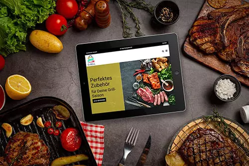 Tablet zeigt den Onlineshop von Q-Sell vor einem Hintergrund mit Grillzubehör und Lebensmitteln