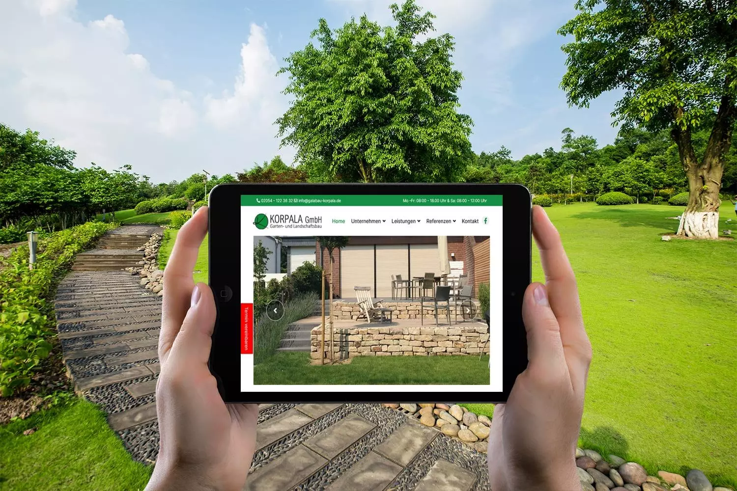 Person hält ein Tablet mit der Joomla Website von Galabau Korpala GmbH, im Hintergrund ein gepflegter Garten