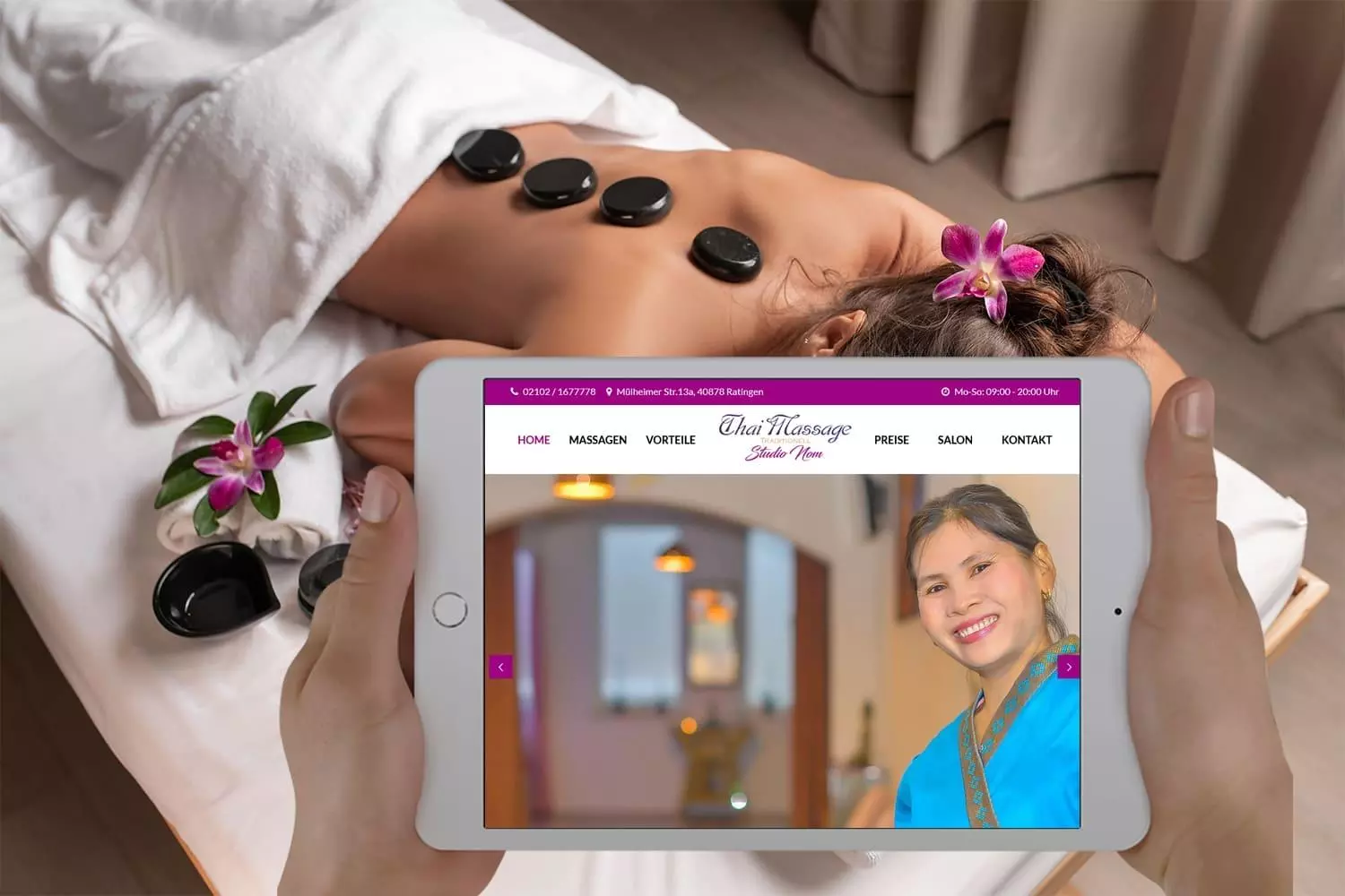 Person hält ein Tablet mit der Thai Massage Nom Webseite in einem Raum, wo eine Frau eine Hot Stone Massage erhält