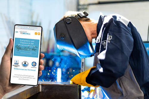 Person hält ein Smartphone mit der Website von Rudi Schindhelm Metall GmbH & Co KG, im Hintergrund ein Schweißer bei der Arbeit