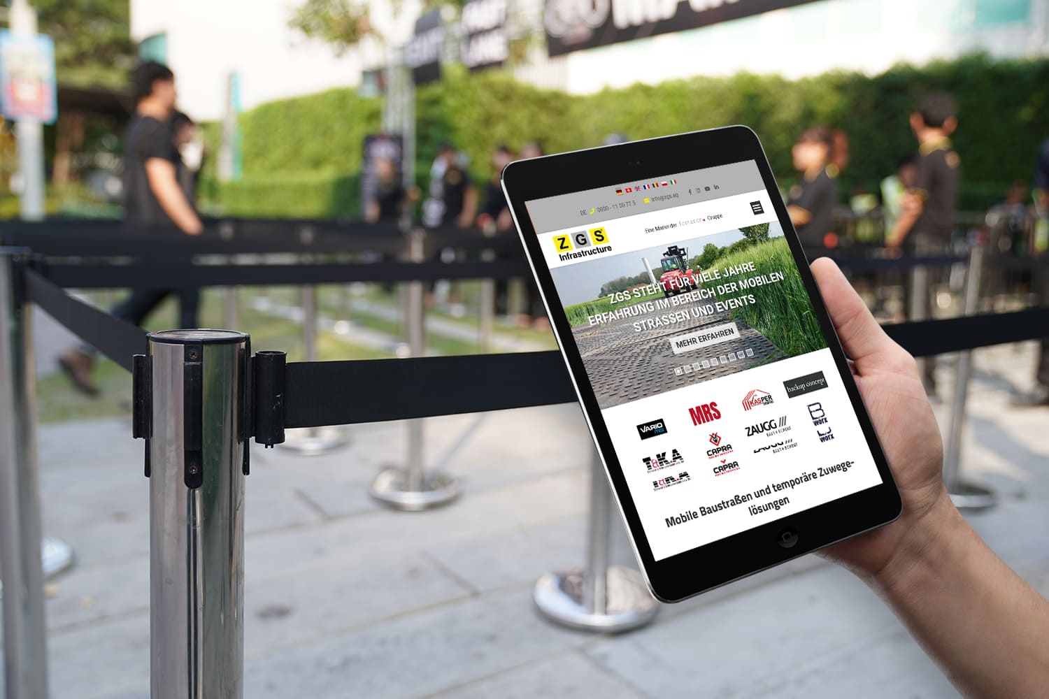 Joomla Webdesign für die ZGS Bau-Website in Düsseldorf – Temporäre Bau- und Eventlösungen online darstellen Person hält ein Tablet mit der Website von ZGS Infrastructure AG, im Hintergrund Absperrbänder für ein Event